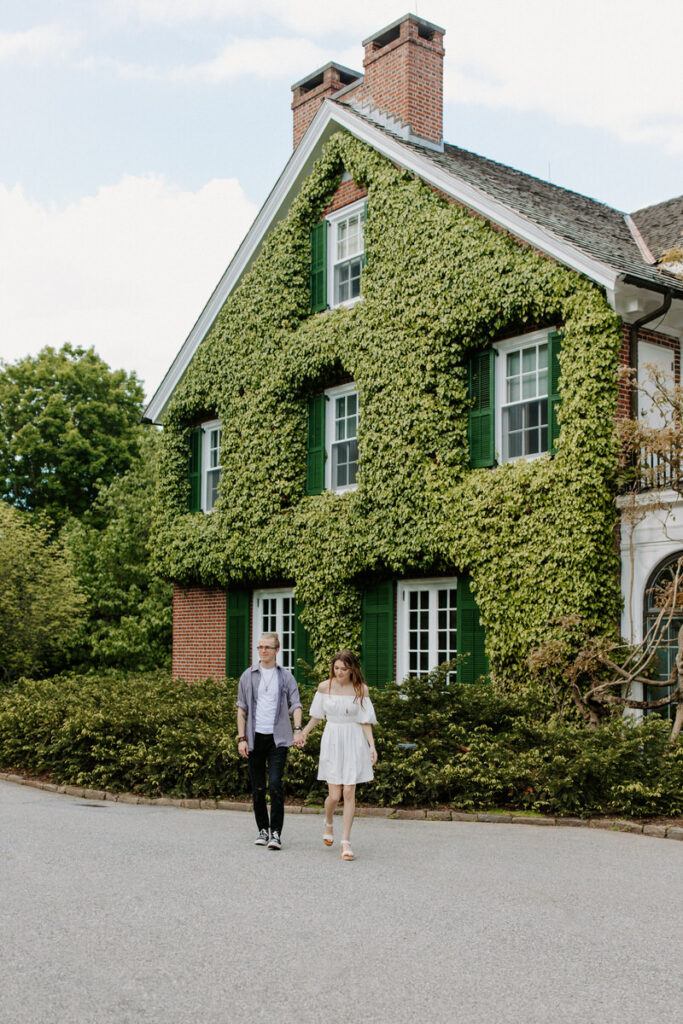 A couple holding hands and walking away from an ivy covered house. 