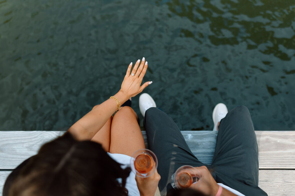 Overhead view of a couple sitting on a pier while one holds out their hand with an engagement ring on it. 