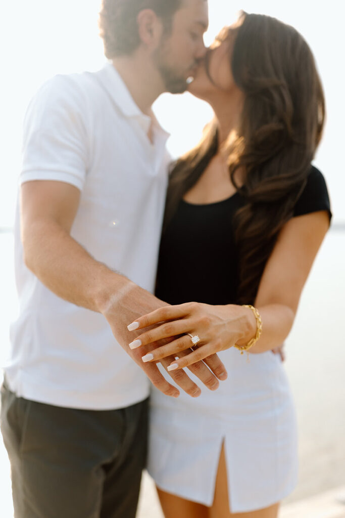A couple holding out a hand with a ring on it as they kiss. 