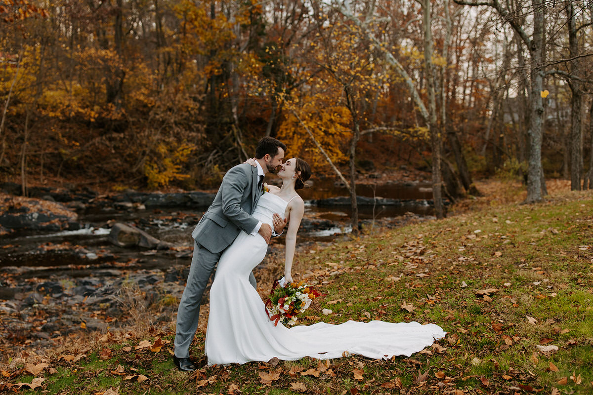 A groom dipping his bride as he leans in for a kiss at one of the best wedding venues in central PA. They are standing on the grassy bank of a creek.
