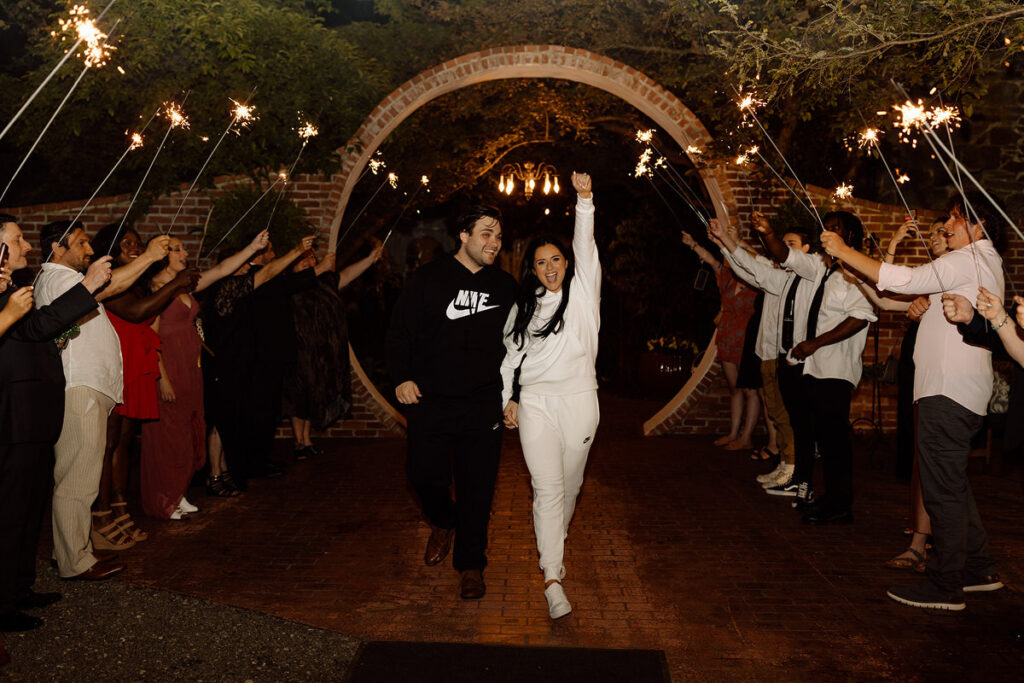 A wedding couple in sweatsuits holding their hands up as they walk through a tunnel of their guests holding up sparklers. 