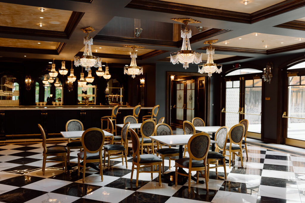 A retro styled room with checkered board floors, small square tables, a bar, and chandeliers hanging from the ceiling. 