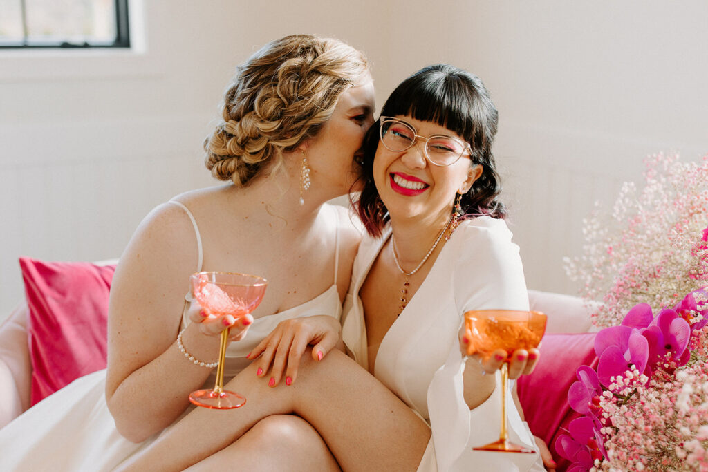 Two brides sitting together on a couch. They are both holding wine glasses and one has their legs over the other. 