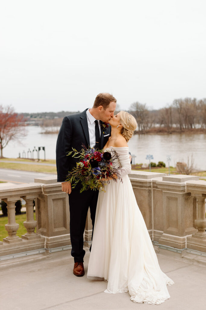 A bride and groom kissing while standing on a balcony that overlooks a lake. 