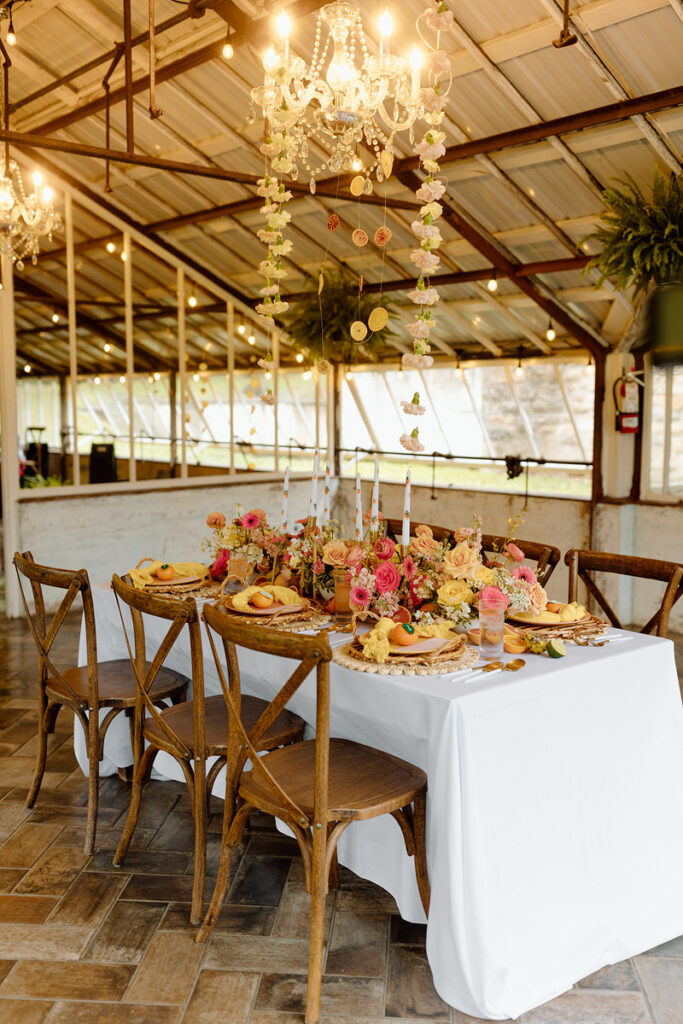 A small rectangular wedding reception table set up in a greenhouse with lights hanging from the ceiling. 