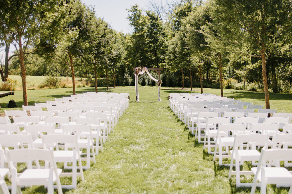 White chairs set up on a lawn with and isle in the middle leading to a small wooden arch. 