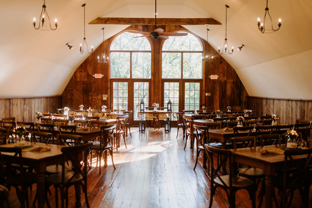 The inside of a barn with large arched windows at the end and wedding reception tables set up along the edges. 