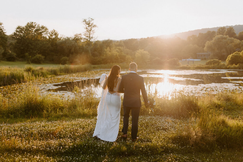 A wedding couple walking in a field towards a small lake as the sun sets. 