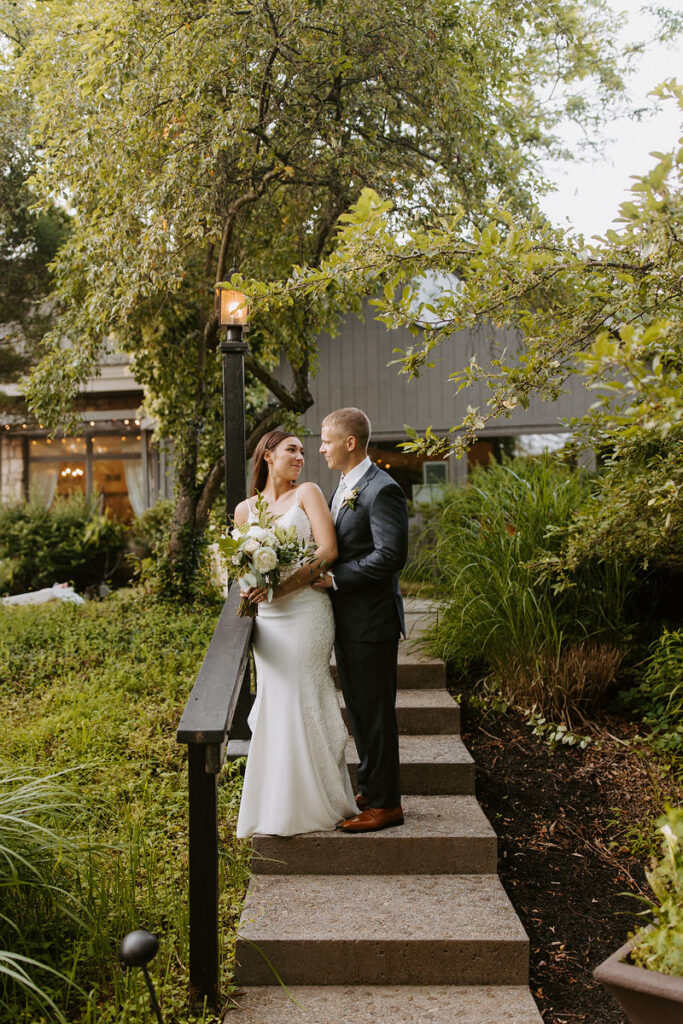 A bride and groom standing on an outdoor staircase together as the groom wraps his arms around her from behind.