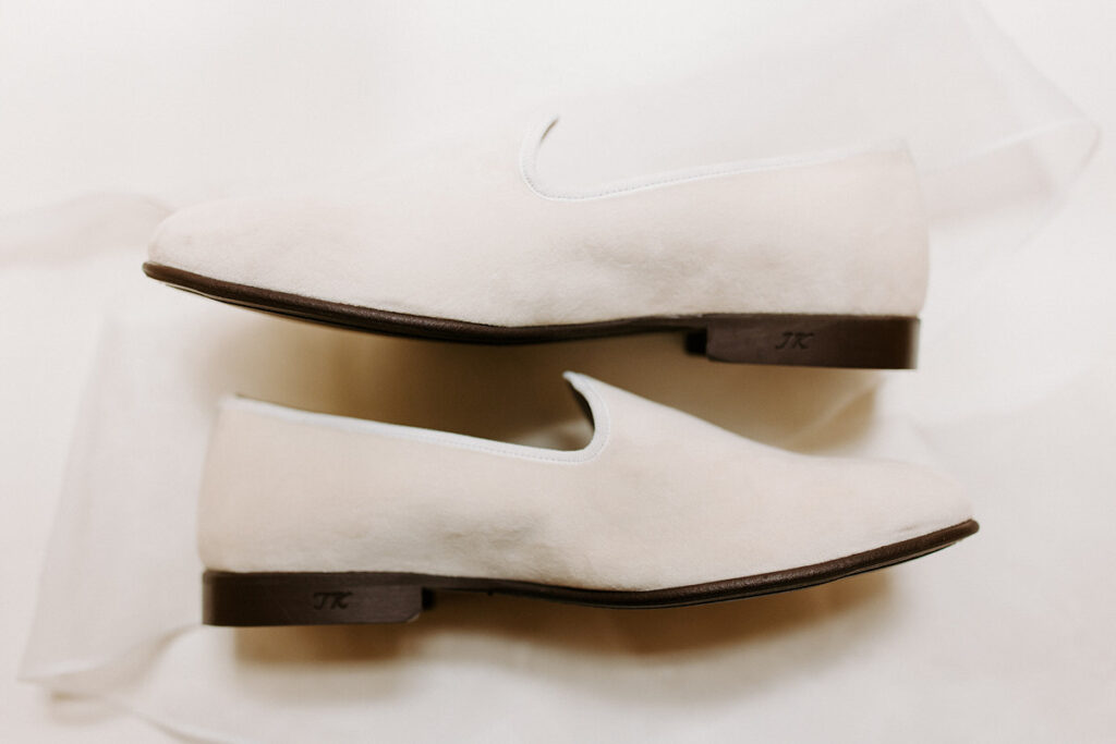 Two white suede shoes with dark soles laying on top of a veil. 