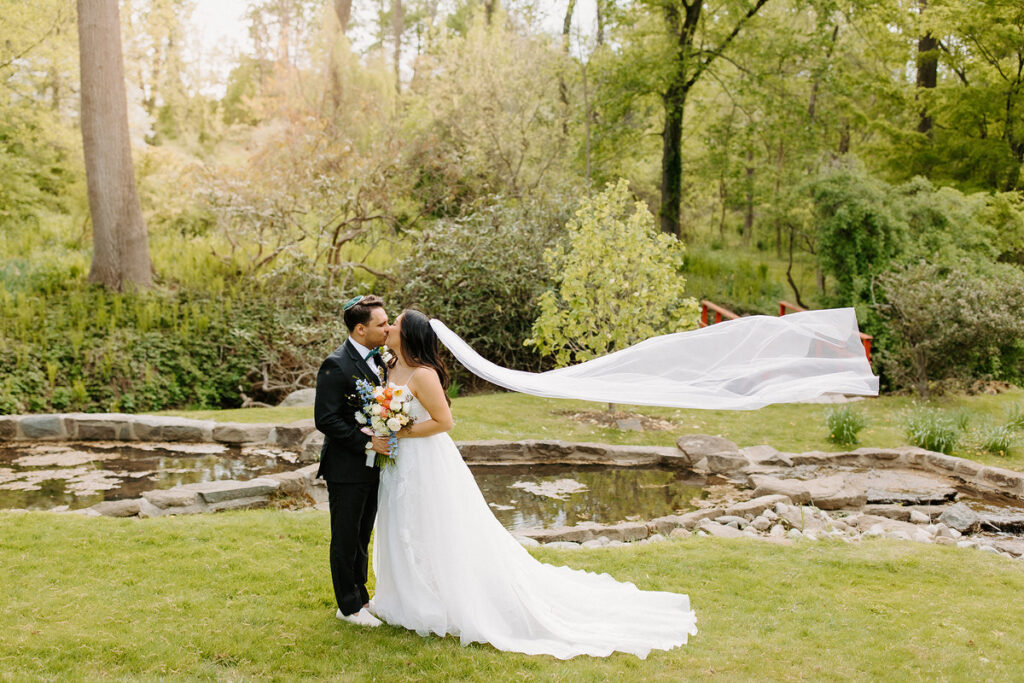 A bride and groom kissing in front of a small pond as the bride's veil floats in the wind. 