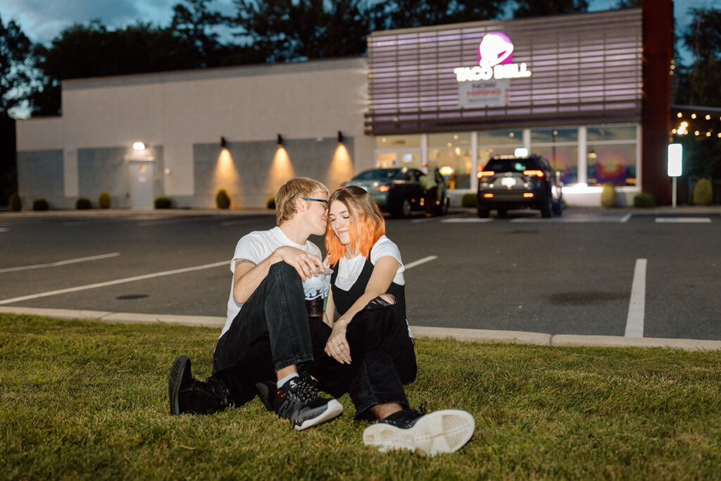 A couple sitting in grass in front of a Taco Bell as one leans in to kiss the other's cheek. 