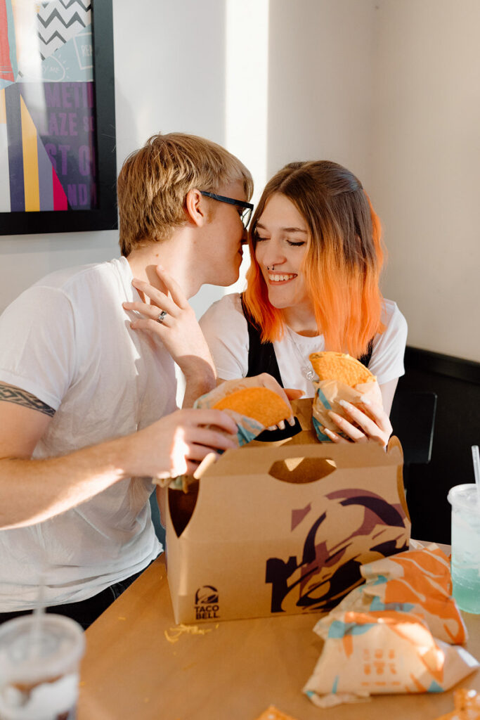A person leaning in to kiss their partner's cheek as they hold tacos. 