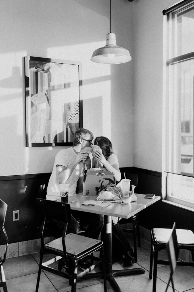A couple in black and white as they kiss in a booth at Taco Bell and hold tacos up to their face. 