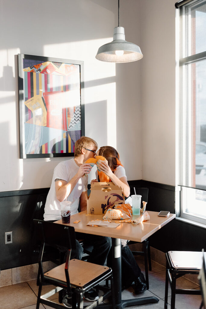 A couple kissing while holding tacos up in front of their faces. 