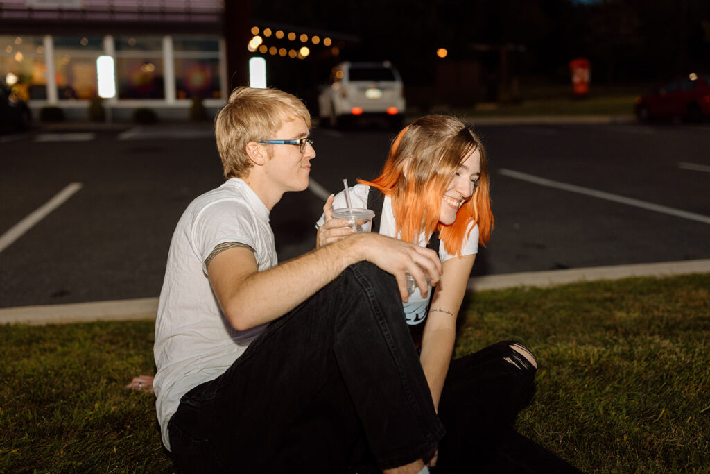 A couple sitting on a grassy patch in a parking lot while laughing. 