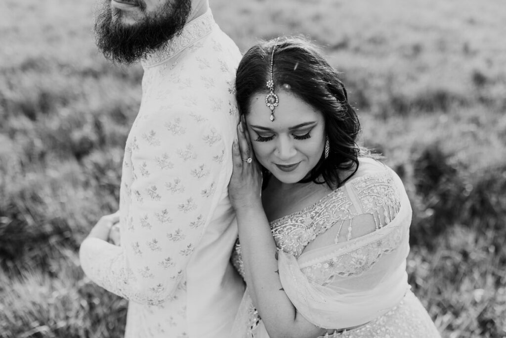 A black and white photo of a couple in a field as the woman leans against the man's back. 