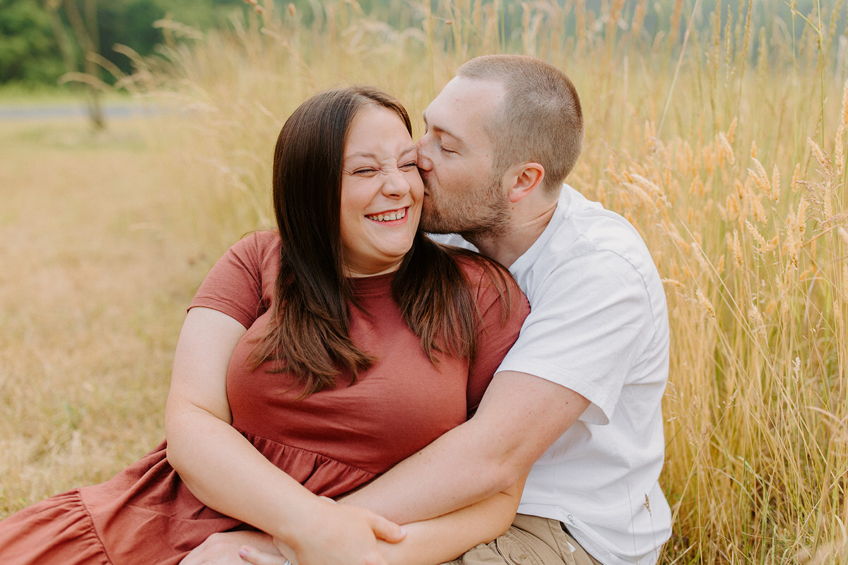 A couple sitting in a field while one kisses the other's cheek as they show off an option of casual photoshoot outfit ideas.