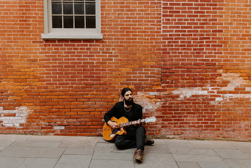 A man sitting on the ground in front of a brick building with a guitar in his lap. 