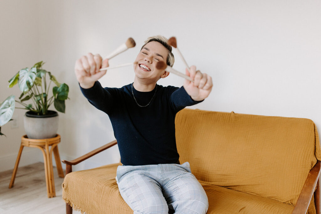 A person sitting on a couch holding out paintbrushes and smiling. 
