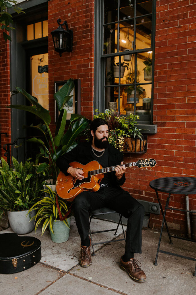 A man sitting at a small table outside with plants behind him as he plays guitar. 
