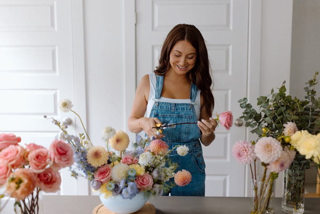 A florist cutting the bottom of a stem with various floral arrangements around her. 
