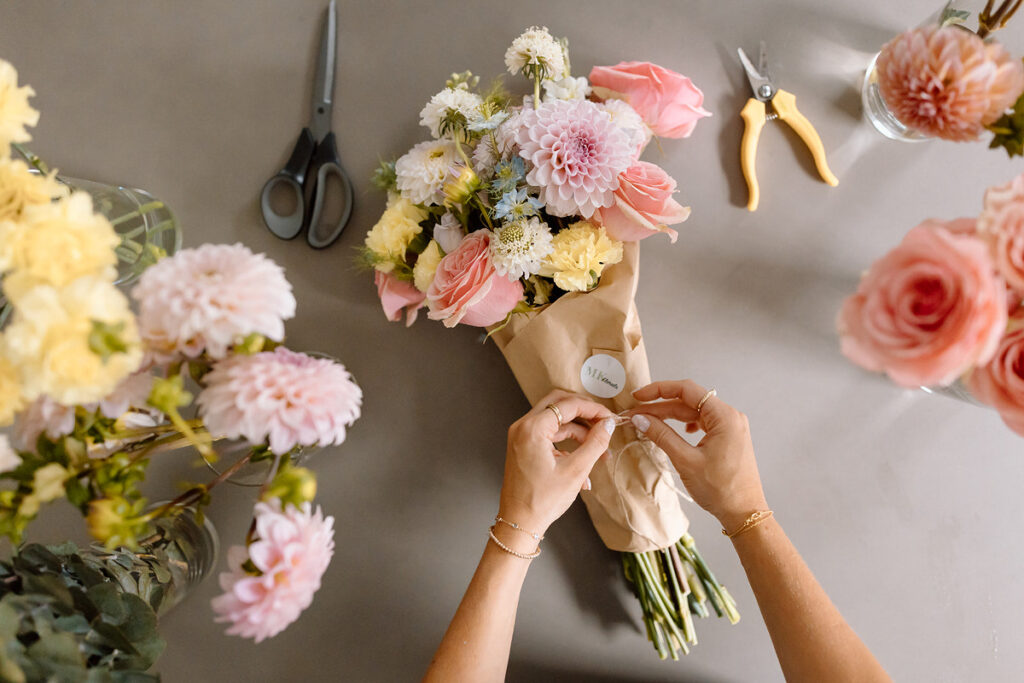 Hands tying paper around a flower bouquet with scissors and pliers laying next to it. 