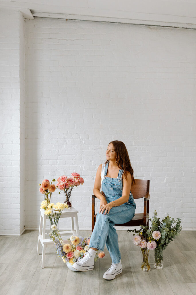 A woman sitting in a chair with vases of flowers around her. 