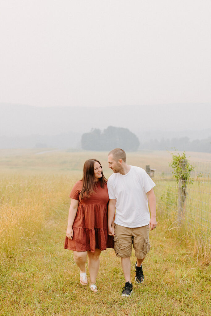 A couple walks hand in hand through a field with tall grass, sharing a glance and a smile in a tranquil, hazy countryside setting.
