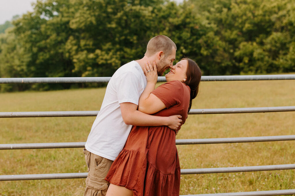 A man in a white shirt embraces a woman in a rust dress by a metal farm gate, sharing a loving kiss in a lush green field