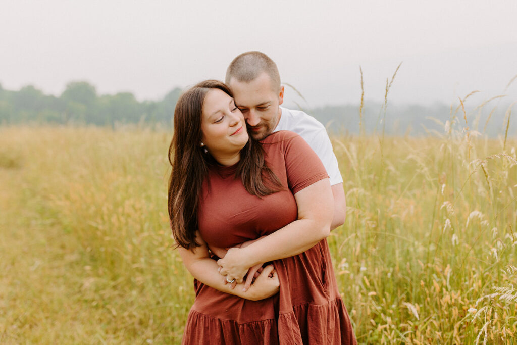 A tender moment as a man hugs a woman from behind in a field, both smiling gently amidst tall golden grass with a soft focus background