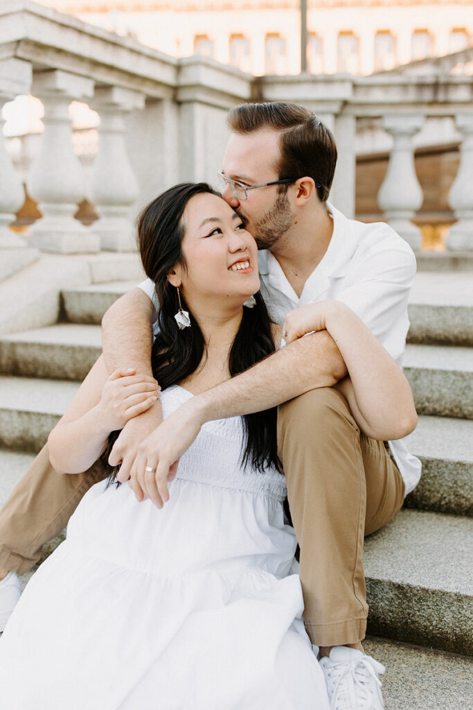 A man in glasses whispers into the ear of a woman in a white dress, both seated on stone steps with a classical architectural backdrop, sharing a tender moment.