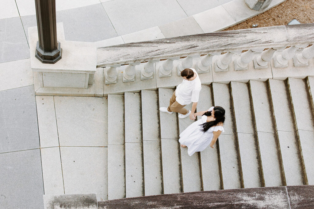 An overhead view of a couple walking down a wide staircase with intricate stone balustrades, holding hands in an urban setting