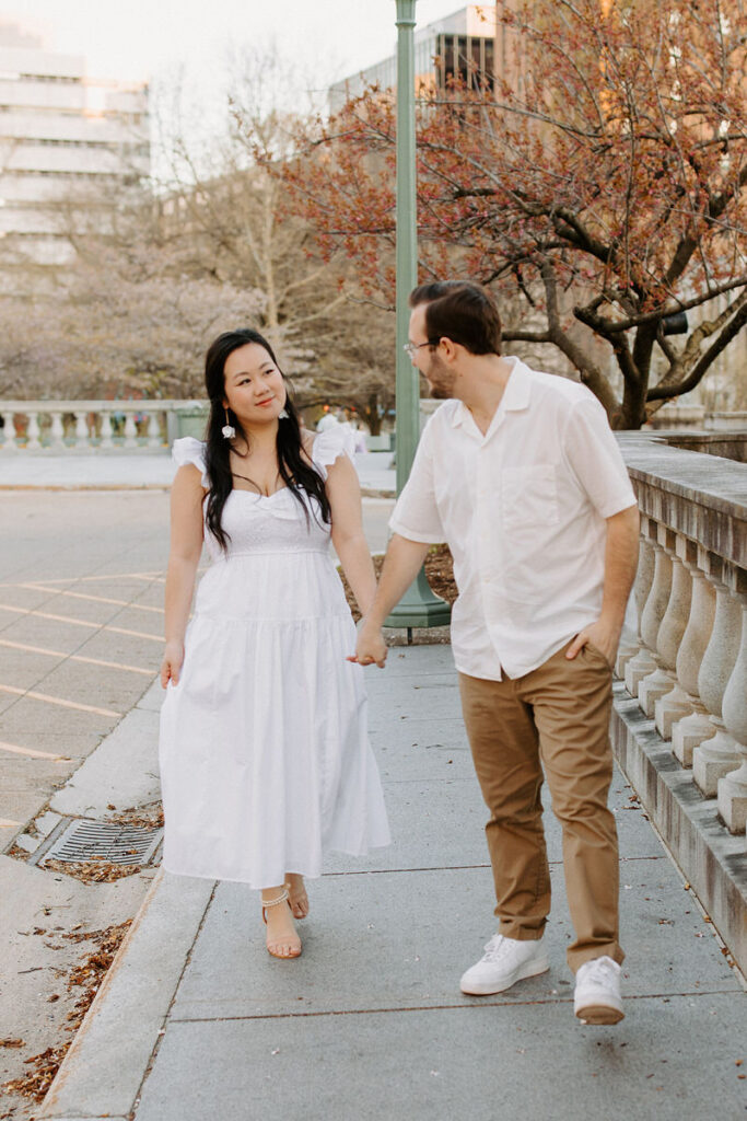 A couple walks side by side along a city walkway, the woman in a white summer dress looking at the man in a white shirt and beige pants, with a balustrade and budding trees in the background