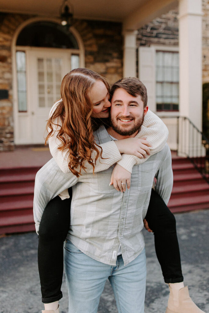 A happy couple in casual wear sharing a piggyback ride and a sweet moment on the steps of a stone-front home with a white door.