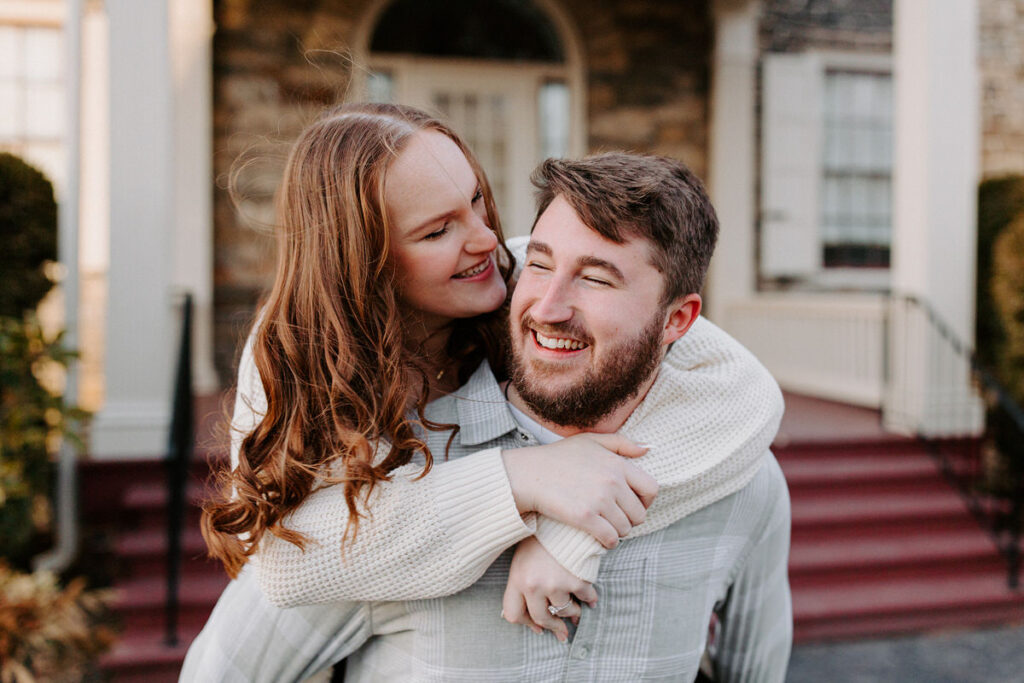 A smiling woman in a cream sweater embracing a bearded man from behind, both looking joyfully at each other in front of a residential backdrop.
