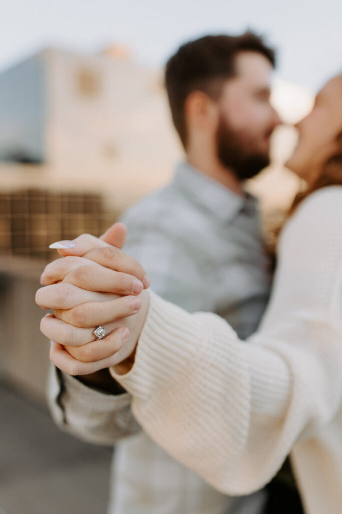 A close-up of a couple's hands intertwined, showcasing an engagement ring, with a soft-focus background of a rooftop setting and the cityscape beyond