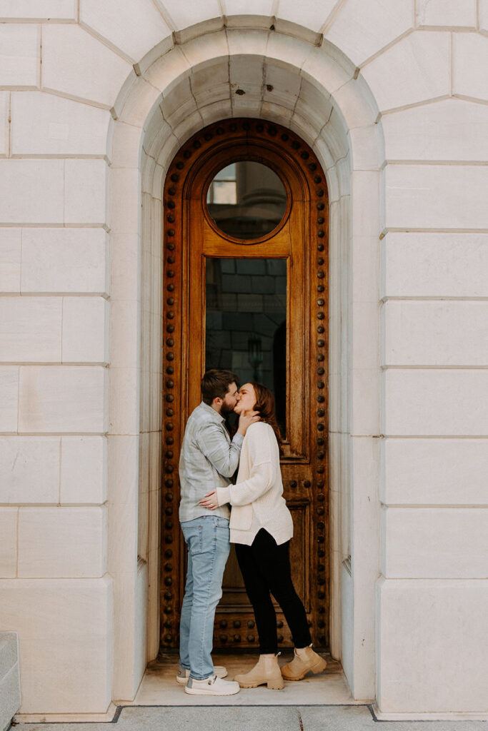 A couple standing in a doorway, sharing a kiss. The arched entryway features a tall wooden door with ornate brass detailing, set in a stone wall.