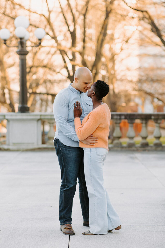 A couple stands in an intimate embrace on a paved walkway. Trees with early spring blossoms and a lamppost line the background in soft focus.