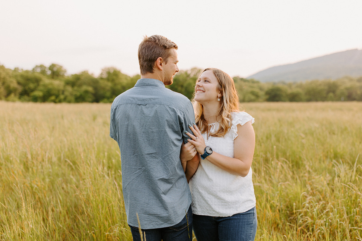 A man and woman in casual clothing stand in a field of tall grass, holding each other and looking into each other’s eyes with affection, with a backdrop of gentle hills in Harrisburg engagement photo