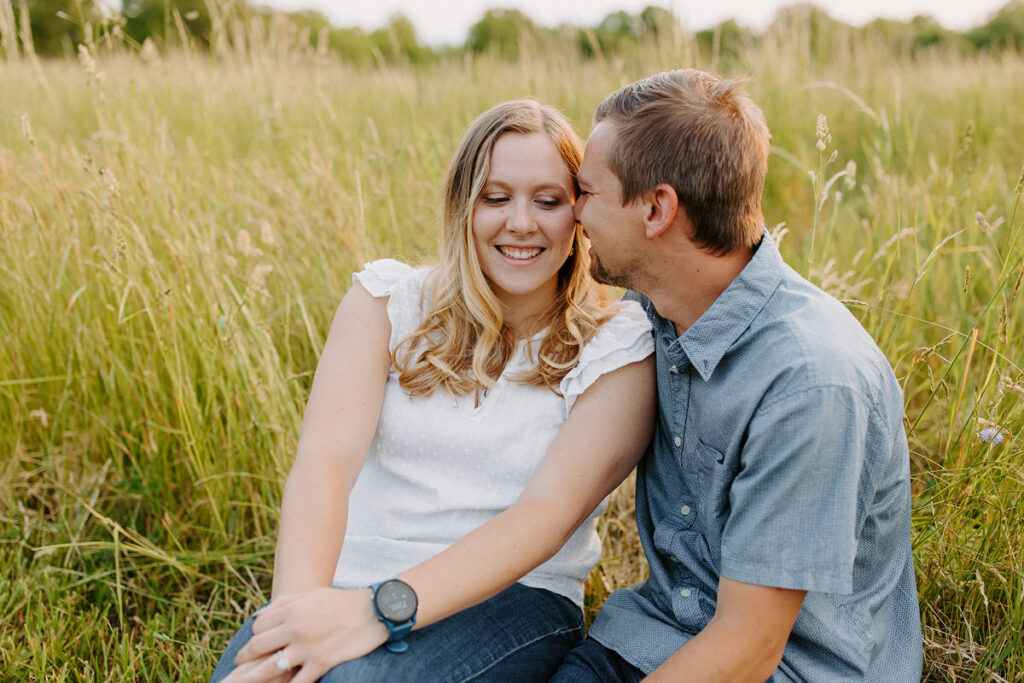 A couple sitting in a field of tall grass as one touches their nose to the other's cheek. 