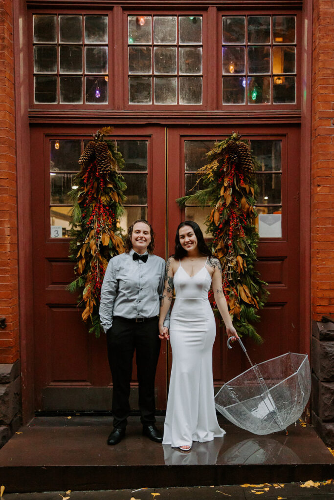 The same couple stands in front of a red door adorned with autumnal wreaths, holding a clear umbrella, smiling towards the camera.