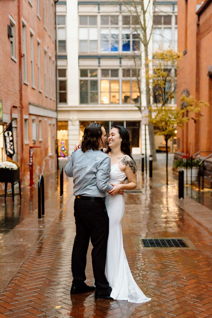 A smiling couple in a rainy alleyway, with the person in a silver shirt embracing the one in a white dress, both gazing affectionately at each other.