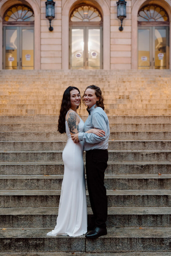 The couple stands on grand stone steps, one in a white dress and the other in a gray shirt, sharing a smile and a loving embrace.