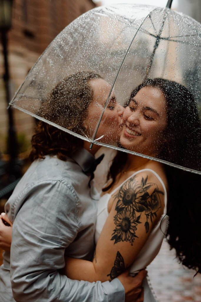 An intimate moment under a transparent umbrella, as one person kisses the other's cheek, both surrounded by raindrops and warmth.