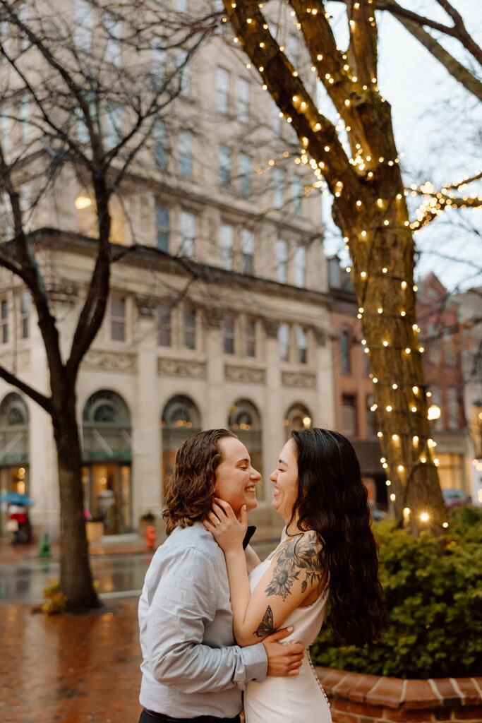 An evening photo capturing the couple embracing under a tree with twinkling lights, creating a romantic city backdrop.
