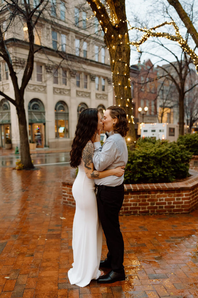 The couple shares a kiss on a rain-kissed street, with a twinkling Christmas tree in the background and the glow of street lights.