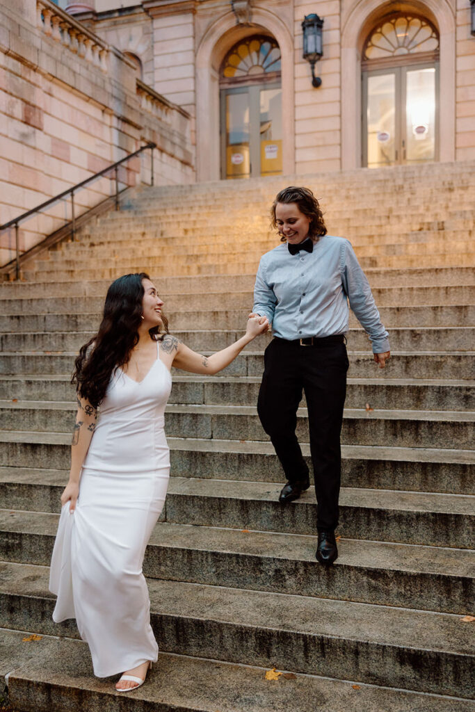 The couple joyfully descends stone steps, holding hands, with the person in the white dress leading and looking back with a smile.