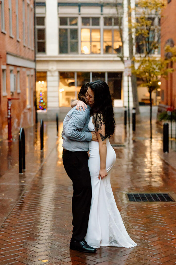 The couple is pictured in a warm embrace in the rain, framed by the brick buildings of a narrow alleyway, sharing a moment of laughter.