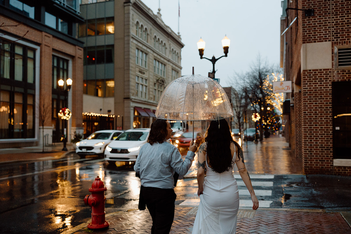 A couple walking away on a wet city street, sharing a clear umbrella with lights reflecting on the ground, creating a romantic urban scene.